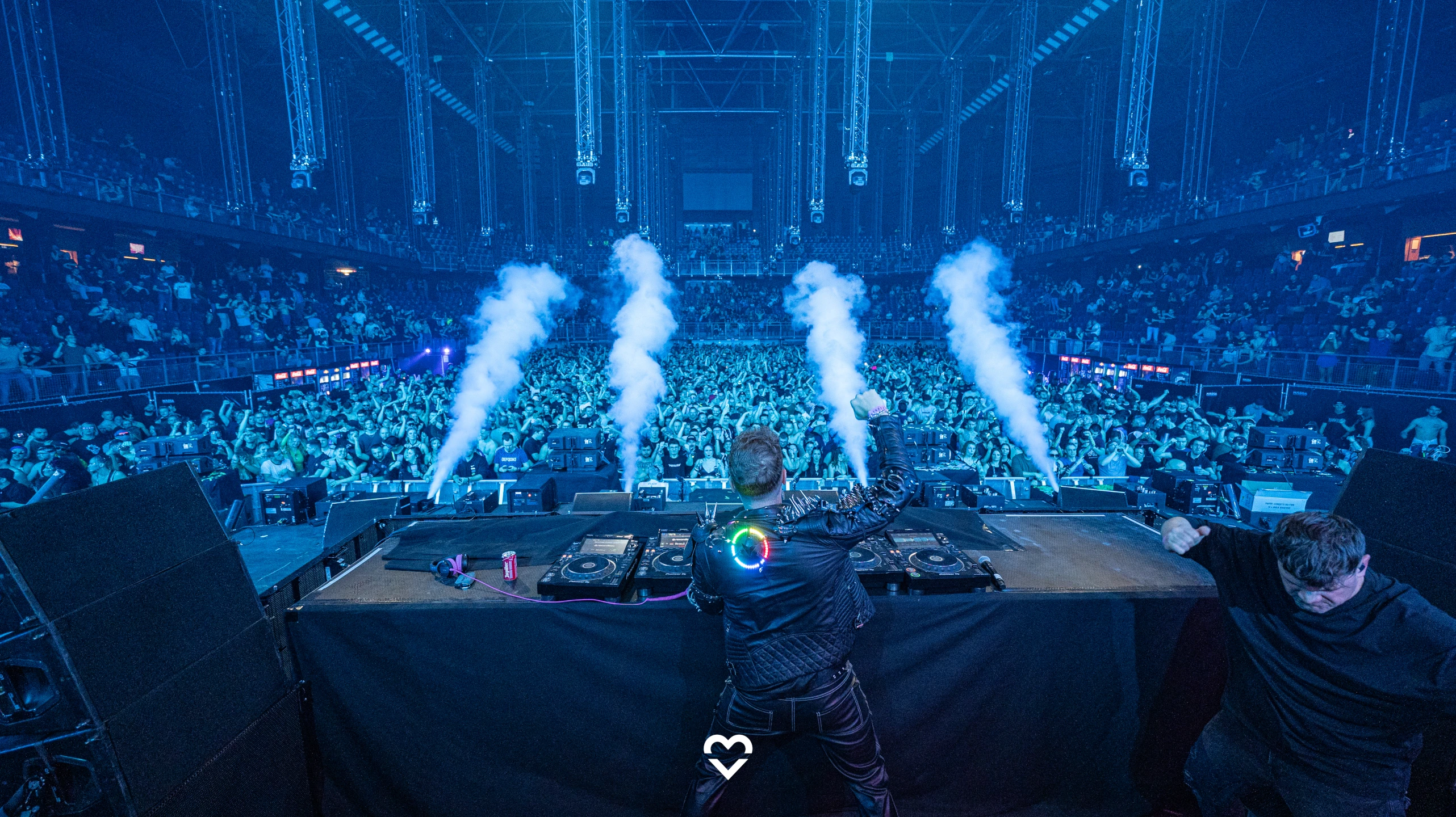 
			Hintergrundbild, das im Vordergrund einen DJ von hinten am Mischpult zeigt,
			der vor einer riesigen Menschenmenge in einer großen Halle Musik spielt.